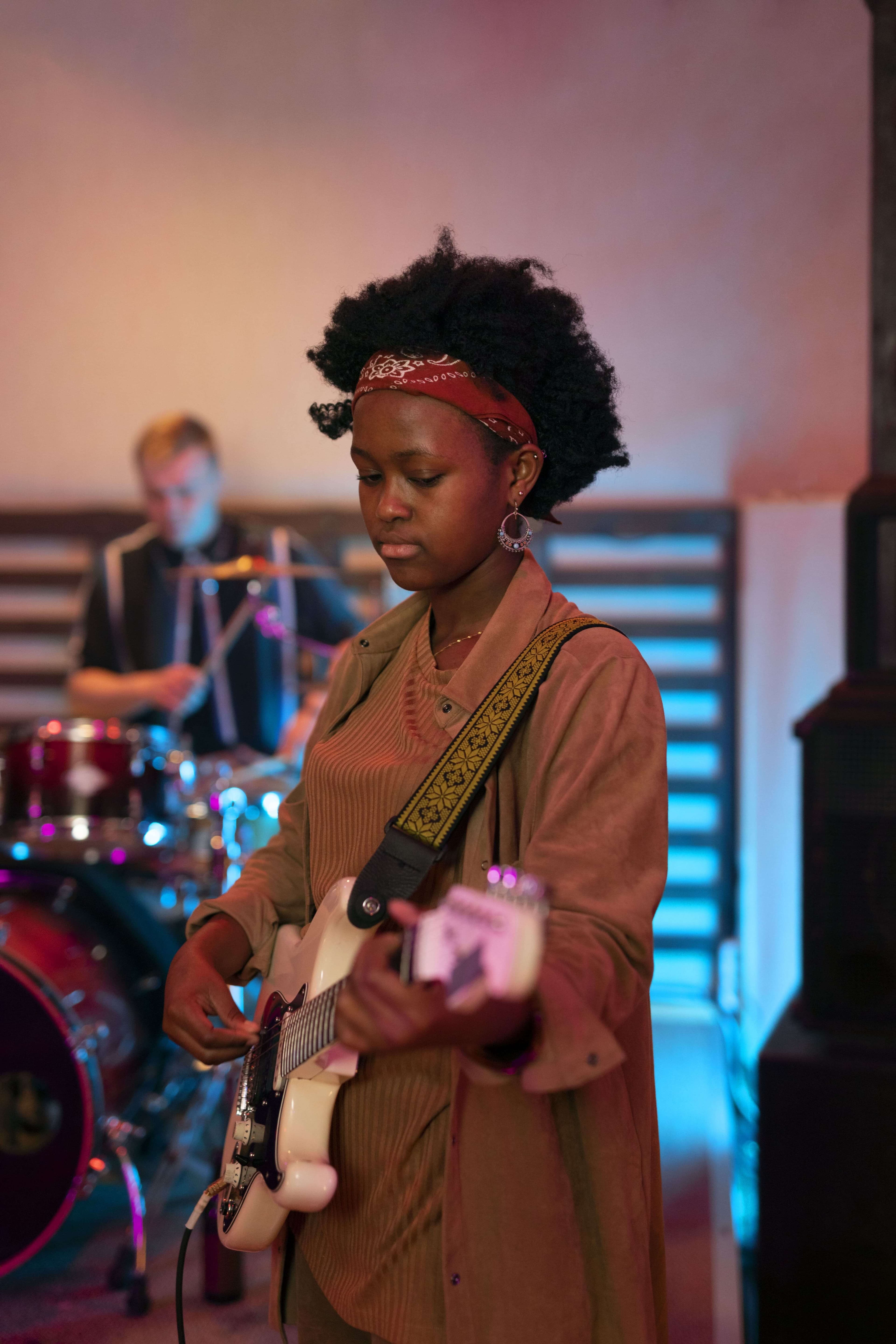 A guitarist performing alongside a drummer in a warmly lit studio.